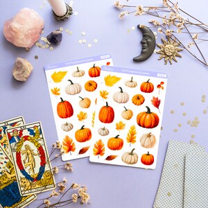 May include: Two sheets of stickers featuring fall-themed pumpkins and leaves. The stickers are on a light purple background with other fall-themed decorations, including a candle, crystals, tarot cards, and a small moon and sun.