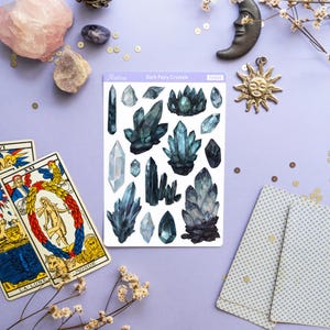 May include: A collection of crystals, tarot cards, and celestial-themed decor on a purple surface. The crystals are in shades of blue and green. The tarot cards are open, and a sticker sheet reads "Dark Fairy Crystals". A candle and a sun pendant are also present.