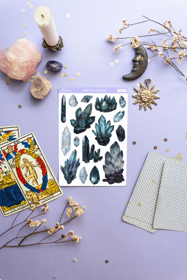 May include: A collection of crystals, tarot cards, and celestial-themed decor on a purple surface. The crystals are in shades of blue and green. The tarot cards are open, and a sticker sheet reads "Dark Fairy Crystals". A candle and a sun pendant are also present.