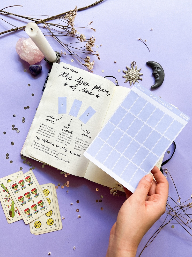 May include: A journal open to a tarot spread layout, with the text "the three phases of time." A hand holds a sheet of blank tarot cards. Tarot cards and decorative items like a candle, crystals, and charms are scattered around.