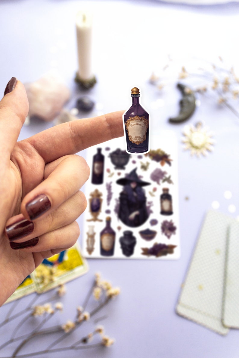 May include: A sticker sheet with a witch, potion bottles, and other magical elements. The sticker sheet is on a white surface and a finger is holding up a potion bottle sticker.