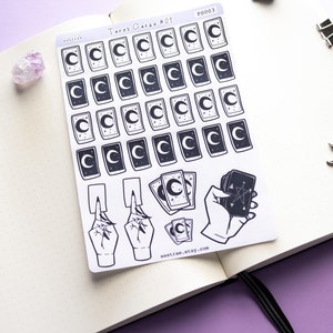 May include: A sheet of stickers featuring tarot card designs with a crescent moon and stars. The stickers are black and white and have a minimalist aesthetic. The sheet also includes a hand holding a tarot card with a pentagram.