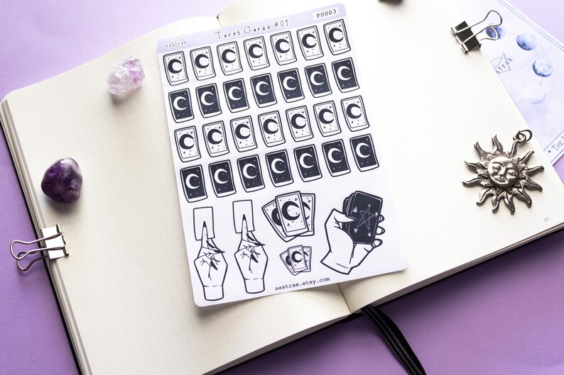 May include: A sheet of stickers featuring tarot card designs with a crescent moon and stars. The stickers are black and white and have a minimalist aesthetic. The sheet also includes a hand holding a tarot card with a pentagram.