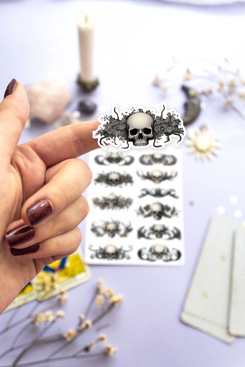 May include: A sheet of stickers featuring a skull with floral accents. The stickers are printed on white paper and are arranged in a grid pattern. The skull is surrounded by black vines and leaves.
