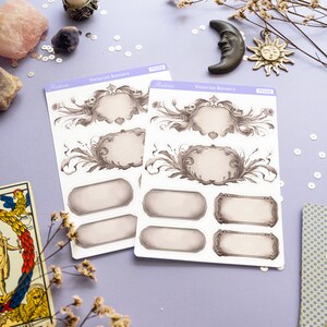 K&ouml;nnte beinhalten: Zwei Bl&auml;tter mit Aufklebern mit viktorianischen Bannern. Die Aufkleber sind wei&szlig; mit grauen und schwarzen Designs. Die Aufkleber befinden sich auf einer violetten Oberfl&auml;che mit wei&szlig;en Pailletten und getrockneten Blumen.