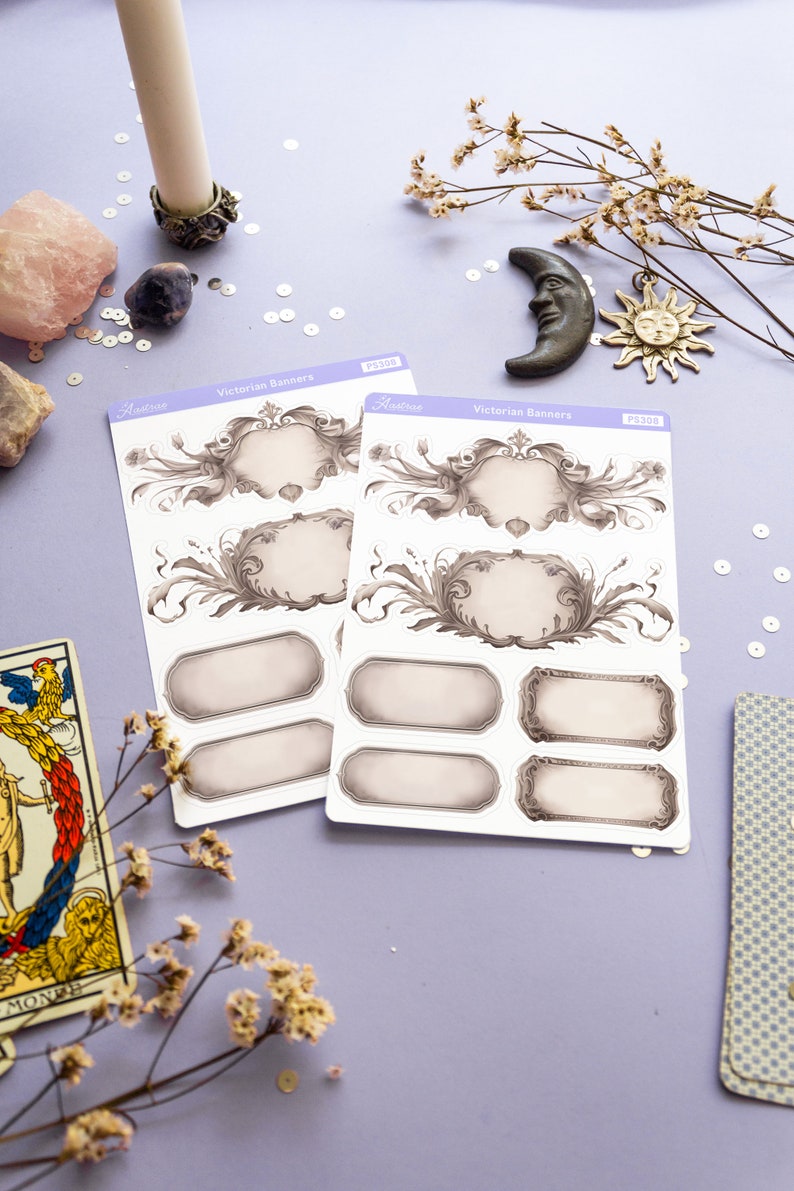 K&ouml;nnte beinhalten: Zwei Bl&auml;tter mit Aufklebern mit viktorianischen Bannern. Die Aufkleber sind wei&szlig; mit grauen und schwarzen Designs. Die Aufkleber befinden sich auf einer violetten Oberfl&auml;che mit wei&szlig;en Pailletten und getrockneten Blumen.