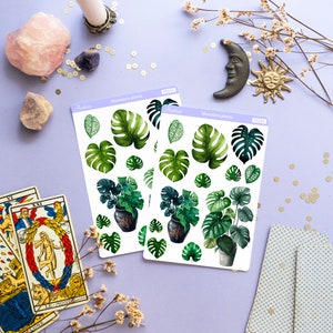 Puede incluir: Dos hojas de pegatinas con ilustraciones de acuarela de plantas de Monstera. Las pegatinas incluyen hojas verdes de varios tama&ntilde;os y plantas en macetas. La imagen tambi&eacute;n incluye cartas del tarot, cristales y objetos decorativos sobre una superficie lavanda.