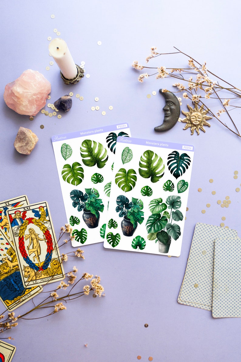 Puede incluir: Dos hojas de pegatinas con ilustraciones de acuarela de plantas de Monstera. Las pegatinas incluyen hojas verdes de varios tama&ntilde;os y plantas en macetas. La imagen tambi&eacute;n incluye cartas del tarot, cristales y objetos decorativos sobre una superficie lavanda.