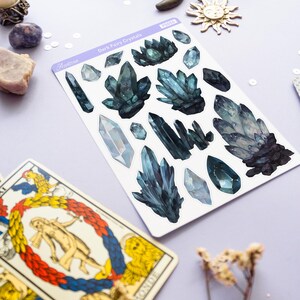 May include: A sheet of stickers featuring dark teal crystal designs, with the text "Dark Fairy Crystals" and the brand name "Austone". Tarot cards and decorative celestial elements, including a crescent moon and sun, are also visible.