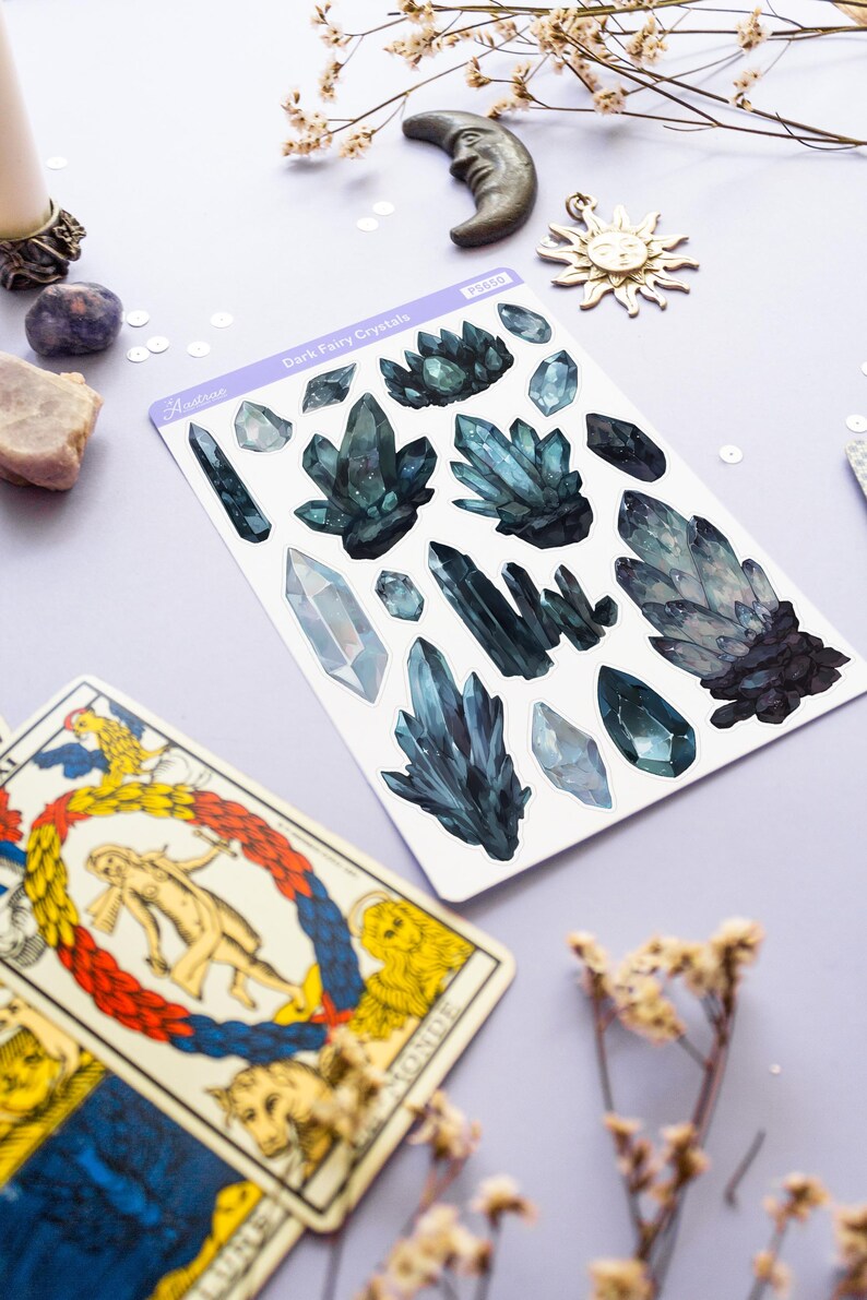 May include: A sheet of stickers featuring dark teal crystal designs, with the text "Dark Fairy Crystals" and the brand name "Austone". Tarot cards and decorative celestial elements, including a crescent moon and sun, are also visible.