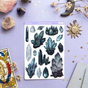 May include: A sheet of stickers labeled "Dark Fairy Crystals" featuring watercolor illustrations of dark blue and teal crystals. The image also includes a tarot card, a sun pendant, and other decorative elements.