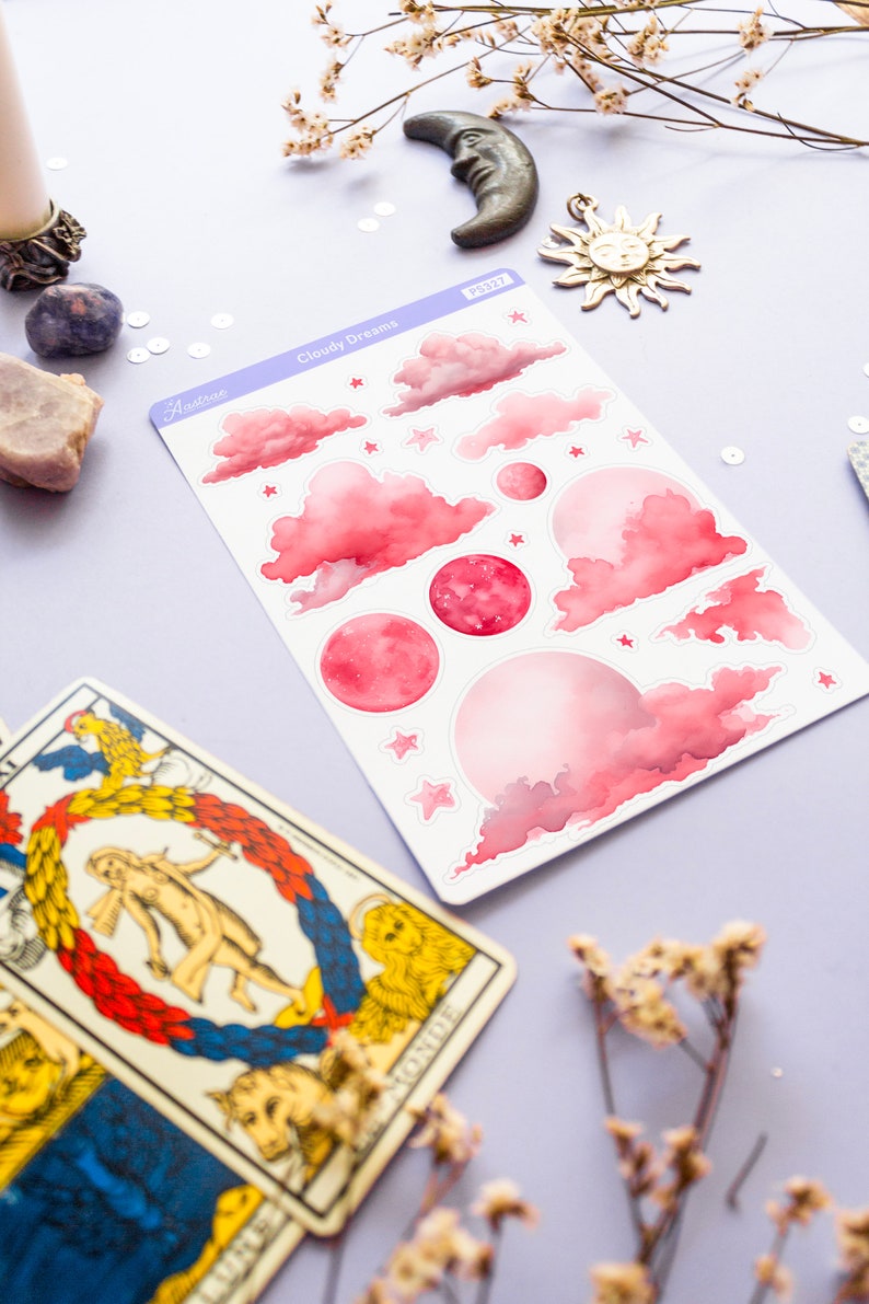 May include: A sheet of pink and white cloudy dream stickers with stars and moons. The stickers are on a purple background with dried flowers and tarot cards.