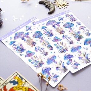 Puede incluir: Dos hojas de pegatinas de hongos iridiscentes. Las pegatinas est&aacute;n sobre un fondo blanco y presentan ilustraciones de acuarela de hongos en tonos morados, azules y rosados. Las pegatinas est&aacute;n etiquetadas como "Hongos iridiscentes" y "Astraea".