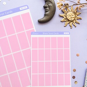 May include: Two sheets of blank pink tarot cards with white borders, labeled "Blank Tarot Cards" and "PS127". A bronze crescent moon and sun charm are placed above the cards. The background is a light purple color with scattered sequins and dried flowers.