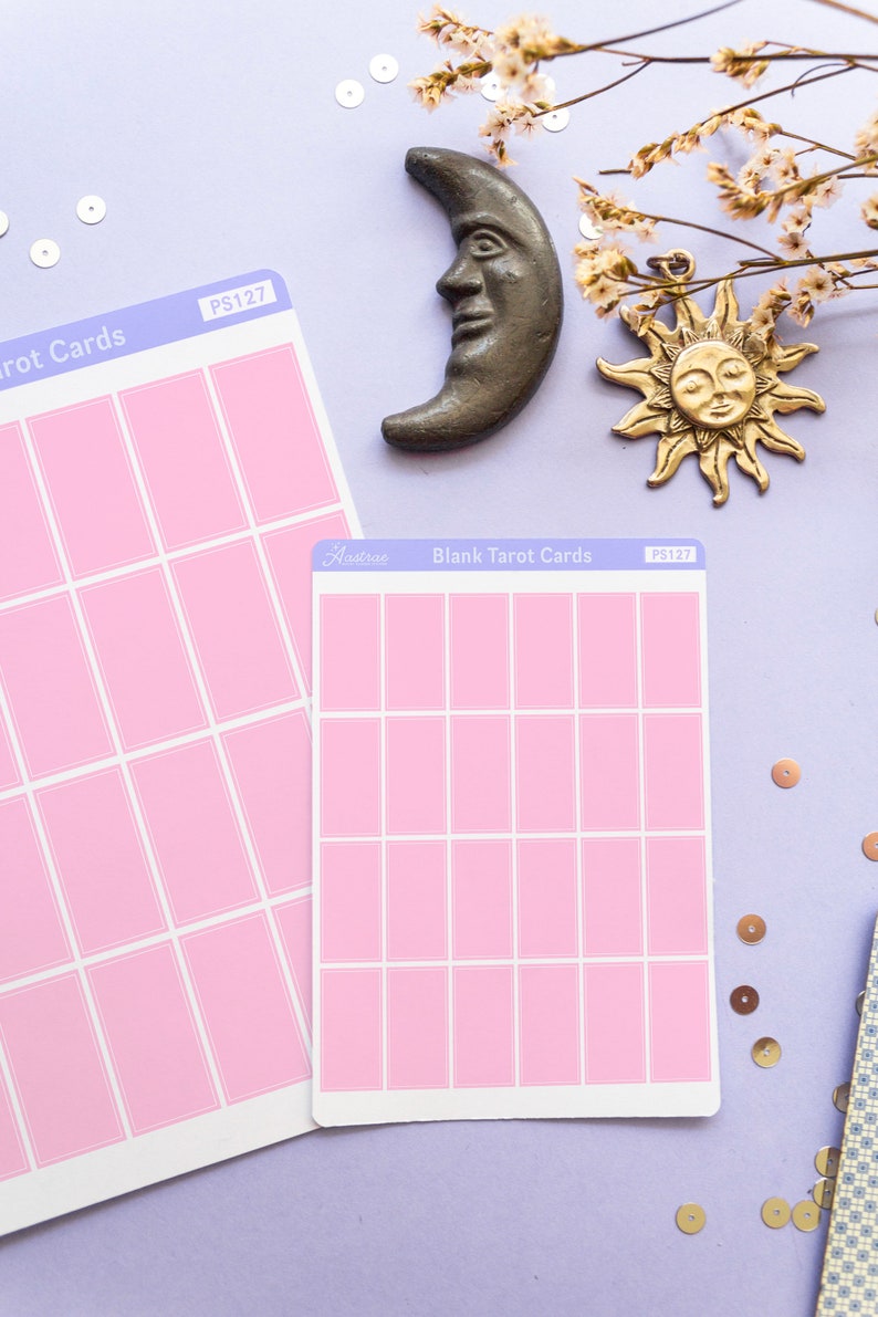 May include: Two sheets of blank pink tarot cards with white borders, labeled "Blank Tarot Cards" and "PS127". A bronze crescent moon and sun charm are placed above the cards. The background is a light purple color with scattered sequins and dried flowers.