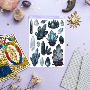 May include: A collection of crystals, tarot cards, and decorative items arranged on a lavender surface. The crystals are various shapes and shades of blue and green. A sticker sheet reads "Dark Fairy Crystals". A candle, moon, and sun charm are also present.
