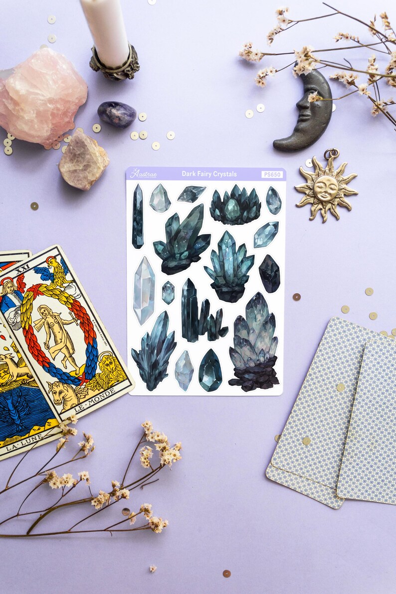 May include: A collection of crystals, tarot cards, and decorative items arranged on a lavender surface. The crystals are various shapes and shades of blue and green. A sticker sheet reads "Dark Fairy Crystals". A candle, moon, and sun charm are also present.