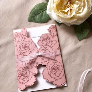 May include: A pink wedding invitation with rose cutouts and a white ribbon. The invitation is open, revealing the inside text. A large, cream-colored rose and green leaves are in the background.