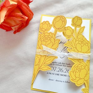 Peut inclure: Une carte d'invitation jaune découpée avec des motifs floraux, avec le texte "07.26.26" et "4 PM AT LAKE SHORE CL". La carte est attachée avec un ruban blanc et une rose en haut à gauche.