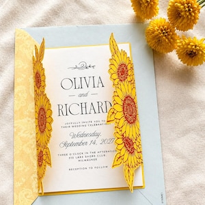 Puede incluir: Una invitación de boda azul claro con detalles de girasoles amarillos. La invitación dice "OLIVIA y RICHARD" e incluye la fecha, hora y lugar de la boda. La invitación está decorada con girasoles amarillos y un sobre con estampado amarillo.