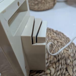 May include: An off-white electric knife sharpener featuring a rectangular plastic housing with two angled slots for blade insertion. A white power cord extends from the side of the unit, which has a smooth, matte finish and a simple, functional design.