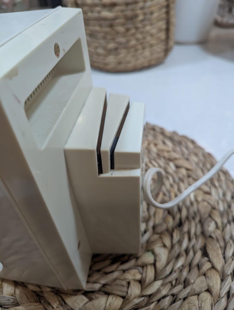 May include: An off-white electric knife sharpener featuring a rectangular plastic housing with two angled slots for blade insertion. A white power cord extends from the side of the unit, which has a smooth, matte finish and a simple, functional design.