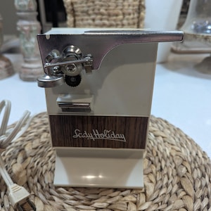 May include: An electric can opener with a cream-colored plastic body and a wood-grain panel featuring the text Lady Holiday in white script. The device includes a metallic cutting mechanism, a black gear, and a white power cord with a plug.