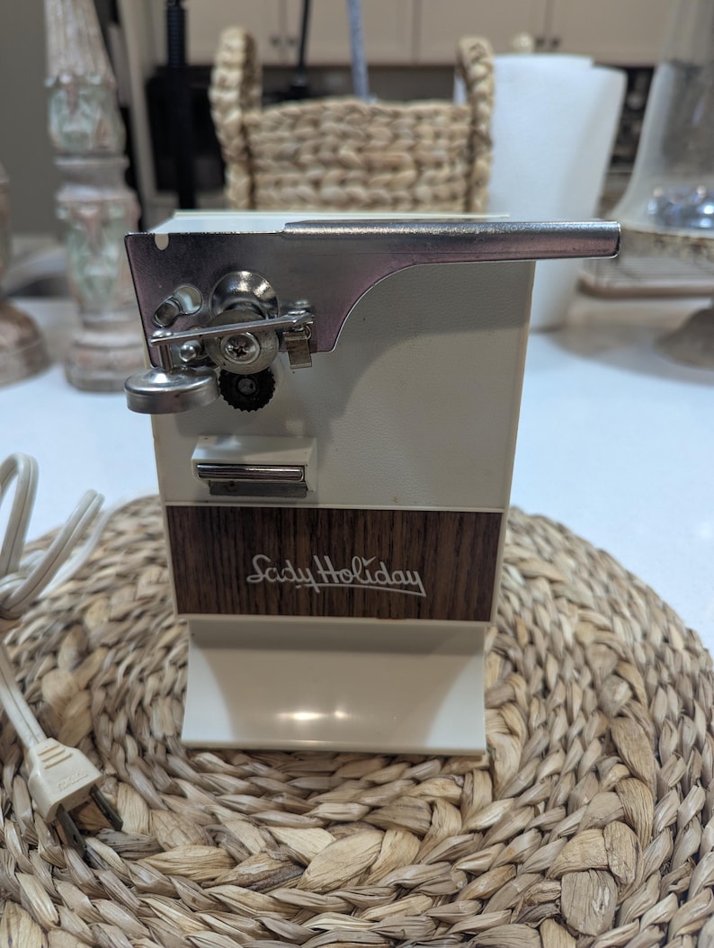 May include: An electric can opener with a cream-colored plastic body and a wood-grain panel featuring the text Lady Holiday in white script. The device includes a metallic cutting mechanism, a black gear, and a white power cord with a plug.