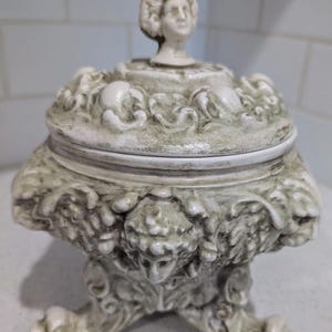 Könnte beinhalten: Eine weiße Keramik-Schmuckdose mit einem Deckel, der eine geformte weibliche Figur mit einer Krone zeigt. Die Dose hat ein detailliertes, verziertes Design mit einer grünen Patina und drei Füßen.