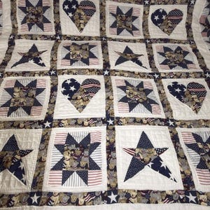 Puede incluir: Una colcha de patchwork con un patrón repetitivo de estrellas y corazones en rojo, blanco y azul. La tela muestra motivos de osos de peluche, secciones a rayas y paneles azul marino con estrellas, separados por bordes acolchados blancos.