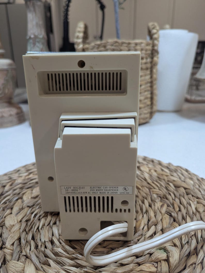 May include: A beige electric can opener and knife sharpener with a white power cord. The back label reads: LADY HOLIDAY 10-0024 ELECTRIC CAN OPENER AND KNIFE SHARPENER 120V 60Hz 1.45A 90W AC ONLY MADE IN JAPAN. The device features ventilation slots.