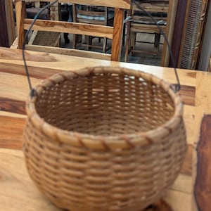 Puede incluir: Una cesta redonda tejida con un asa de madera y un marco de alambre metálico. La cesta está hecha de mimbre marrón claro y tiene un diseño sencillo y rústico. El asa es de madera marrón oscuro.