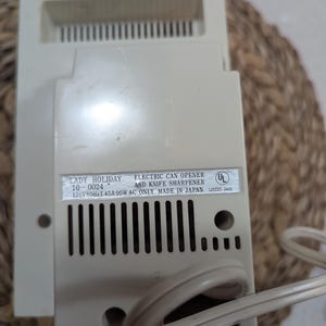 May include: An off-white Lady Holiday electric can opener and knife sharpener with a power cord. The label reads: LADY HOLIDAY 10-0024 ELECTRIC CAN OPENER AND KNIFE SHARPENER 120V 60Hz 1.45A 90W AC ONLY MADE IN JAPAN UL LISTED 243E. It has vertical vents.