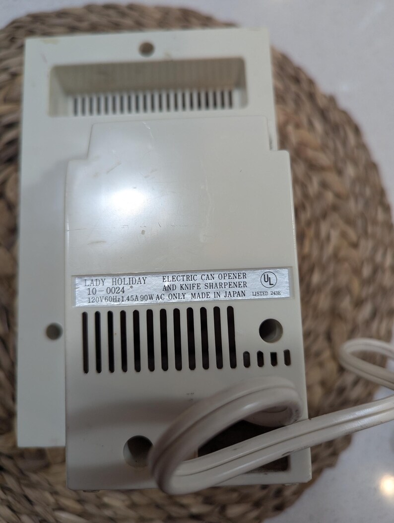 May include: An off-white Lady Holiday electric can opener and knife sharpener with a power cord. The label reads: LADY HOLIDAY 10-0024 ELECTRIC CAN OPENER AND KNIFE SHARPENER 120V 60Hz 1.45A 90W AC ONLY MADE IN JAPAN UL LISTED 243E. It has vertical vents.
