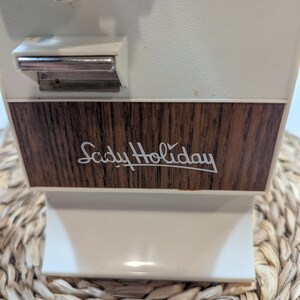 May include: An electric can opener with a white plastic body and a dark brown wood-grain panel on the front. The text "Lady Holiday" is printed in white script on the wood-grain section. A silver metal lever is positioned above the panel.