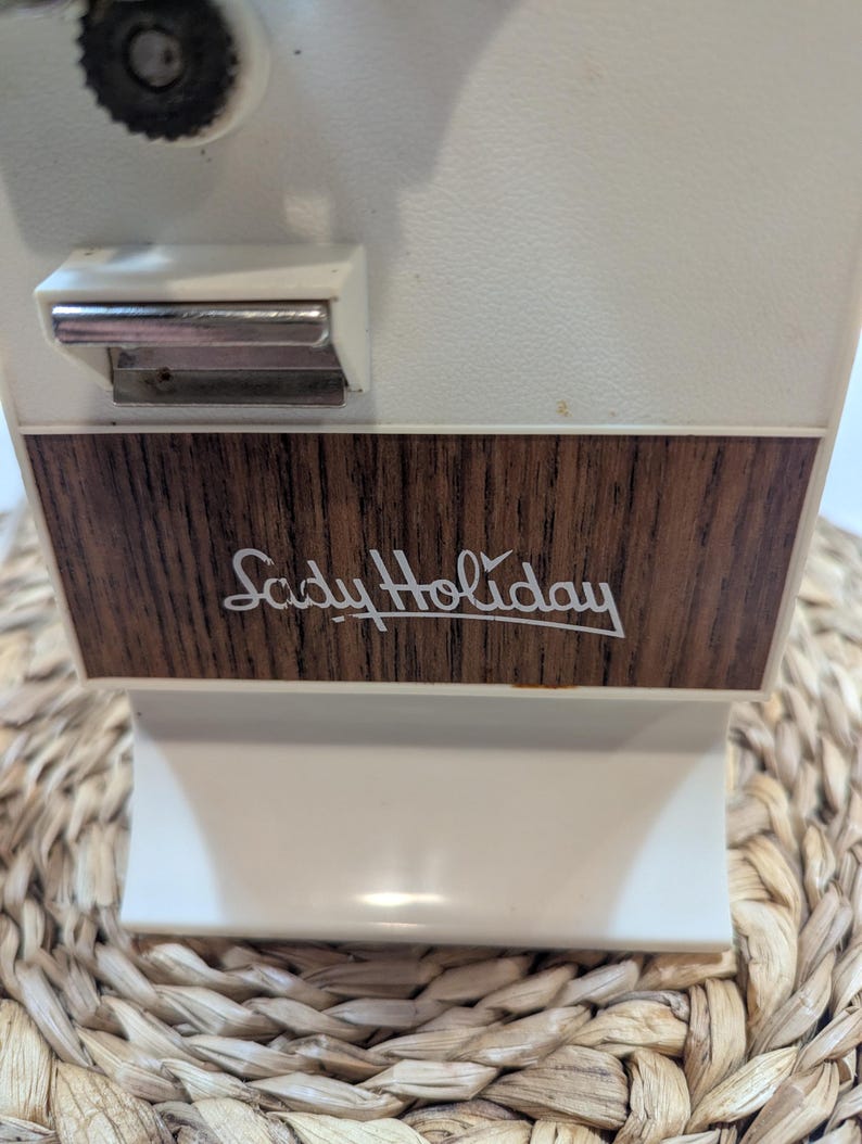 May include: An electric can opener with a white plastic body and a dark brown wood-grain panel on the front. The text "Lady Holiday" is printed in white script on the wood-grain section. A silver metal lever is positioned above the panel.
