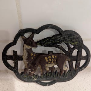 Pode incluir: Placa decorativa em ferro fundido com um veado e um veado em uma cena rústica. O veado é marrom com manchas amarelas, em frente a uma cerca e uma árvore. A placa tem um acabamento escuro e envelhecido, adequado para decoração de casa.