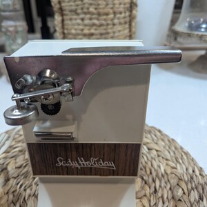 May include: An electric can opener with a white plastic body and a wood-grain panel featuring the text Lady Holiday in cursive script. The device includes a silver metal cutting mechanism with a lever and a small circular knob on the front side.