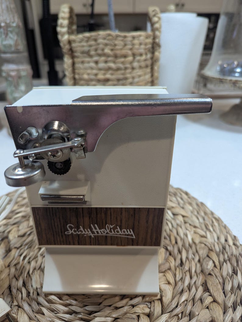 May include: An electric can opener with a white plastic body and a wood-grain panel featuring the text Lady Holiday in cursive script. The device includes a silver metal cutting mechanism with a lever and a small circular knob on the front side.
