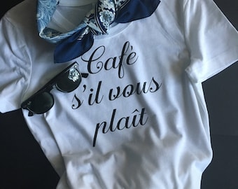May include: White t-shirt with the text "Cafe s'il vous plait" in black cursive lettering. A blue and white patterned bandana is tied around the neck of the shirt. Sunglasses are placed on top of the shirt.