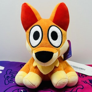 May include: A plush toy shaped like a dog with orange fur, large white eyes with black pupils, and a black nose. The ears have dark red inner sections, and the chest area is light yellow. A purple tag is attached to the side of the head.