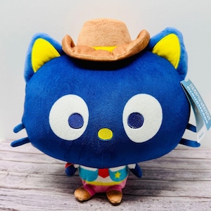 May include: A plush toy of a blue cat character wearing a tan cowboy hat. The cat has large white eyes with dark blue pupils, yellow inner ears, and a small yellow nose. It wears a white, red, and blue vest with a yellow star and pink pants.