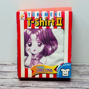 Peut inclure: Un t-shirt blanc avec un graphique de personnage d'anime aux cheveux violets, emballé dans une boîte en carton à rayures rouges et blanches. La boîte affiche le texte TOKIMEKI T-Shirt II, le logo Konami et des caractères japonais sur une bannière jaune.