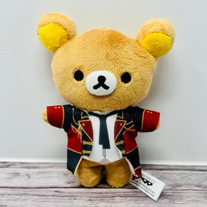 May include: A plush bear toy with a brown body, yellow ears, and black eyes and nose. The bear is wearing a red and black jacket with gold accents and a white shirt and black tie. A small tag is attached.