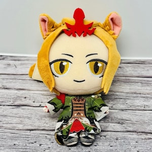 May include: A plush doll with a tan face, yellow eyes, and a small smile. The doll has yellow hair with red accents and wears a green and red outfit. The doll is approximately 6 inches tall.