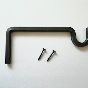 May include: A black, metal curtain rod bracket with a curved end and two black screws. The bracket is L-shaped, designed to hold a curtain rod. The screws are for mounting the bracket to a wall.