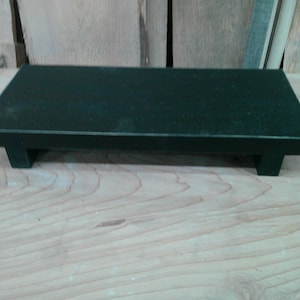 May include: A rectangular, dark green wooden serving tray with a flat surface and short, T-shaped legs. The tray is set on a light-colored wooden surface, with a natural wood grain pattern visible.