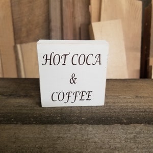 May include: White wooden sign with black text that reads "HOT COCA & COFFEE".