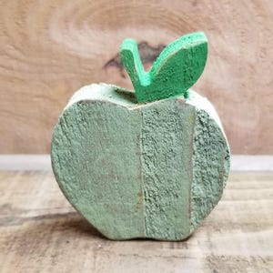 May include: A green wooden apple with a green leaf on top. The apple is made of wood and has a rustic look.
