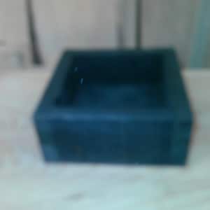 May include: A square, dark blue wooden box with a hollow center and thick, solid walls. The container features a smooth, matte finish and a simple, geometric construction with visible joinery details at the corners of the frame.